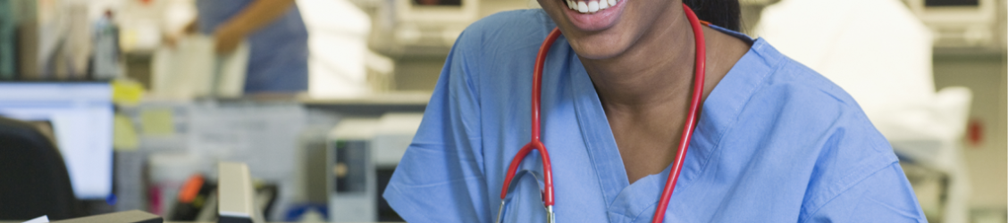nurse in blue scrubs sits at nursing station, writing on clipboard