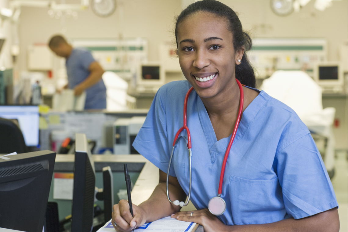 nurse in blue scrubs sits at nursing station, writing on clipboard